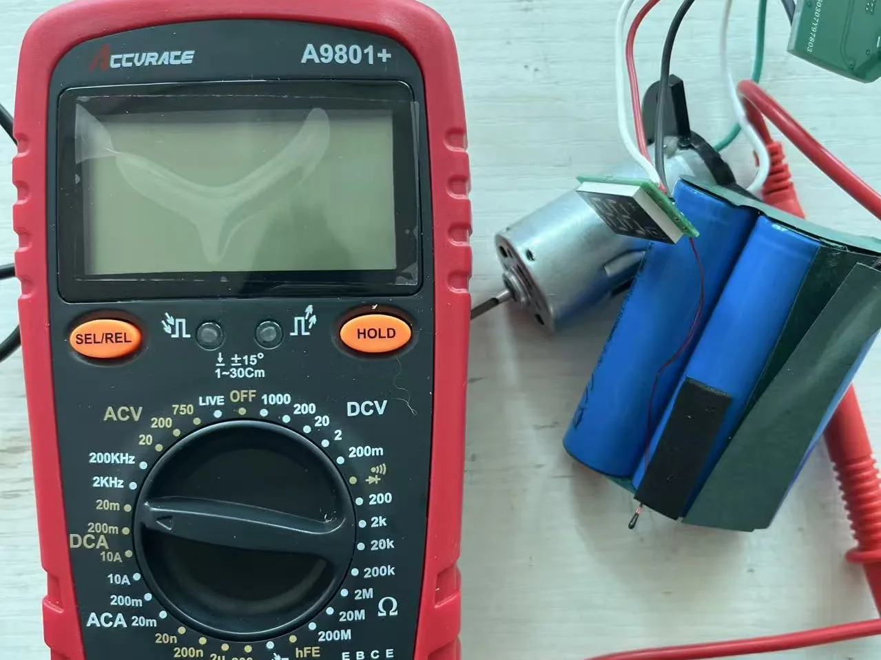 Multimeter on the workbench next to a DC pump motor and a lithium battery pack with its protection board